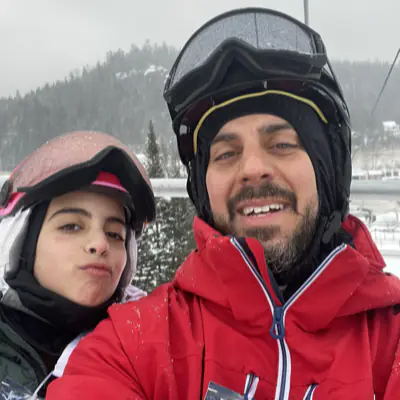 Mika and her dad on a chairlift in helmets and goggles, snow on their jackets.